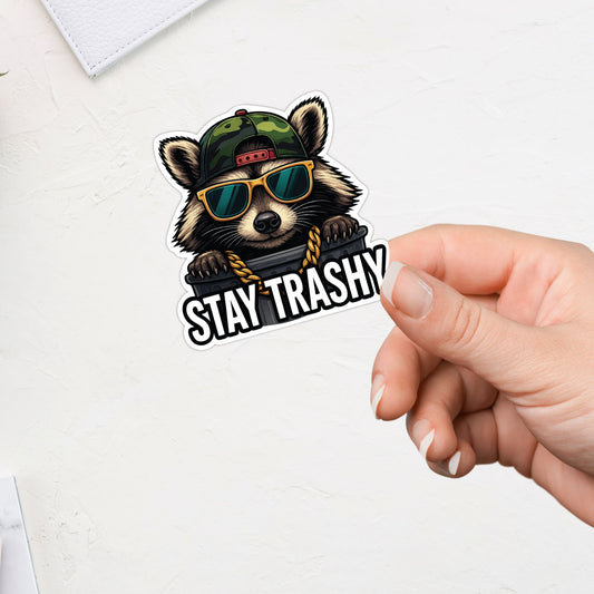Sticker with a raccoon wearing sunglasses and a cap, with 'Stay Trashy' text, held by a hand on a white background.