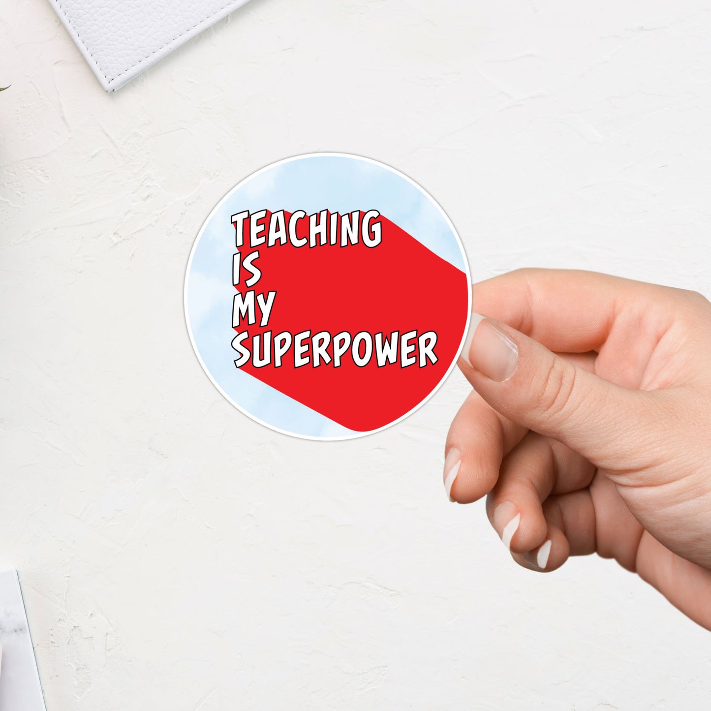 Hand holding a circular sticker with 'Teaching is my superpower' text on a light background